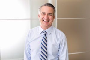 older man smiling and wearing light blue dress shirt and blue striped tie 